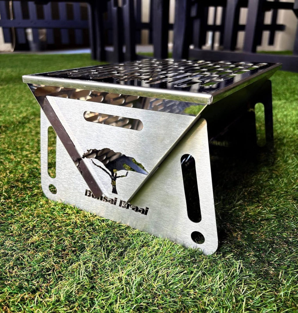 The Bonsai Braai set up on grass, showing the brushed 430 stainless steel sides with a thorn tree cut out and the grid on top.