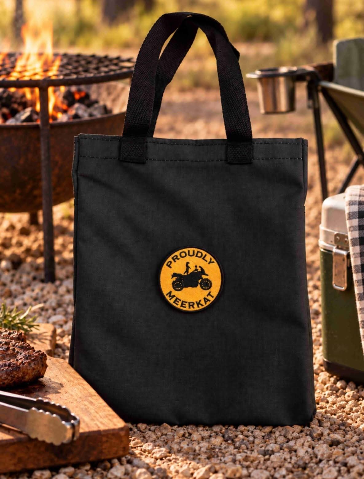 The Proudly Meerkat ripstop canvas carry bag with its embroidered badge, standing beside a fire.